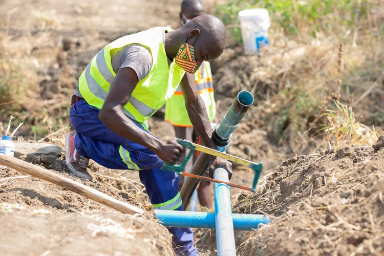 Technicians in high-visibility vests join sections of blue pipe in a trench in Mangochi District, Malawi