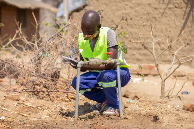 A water technician in a high-visibility vest records readings at a tap stand in Mangochi District, Malawi